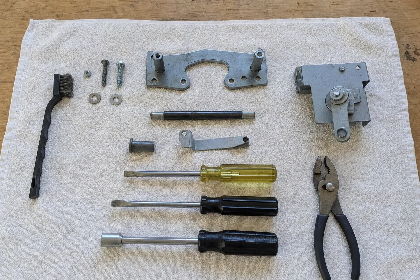 Tools laid out on a white cloth with a spring-loaded hinge mechanism, a short steel shaft, a bushing, a lever link, a twin-lobe bracket and loose screws and washers